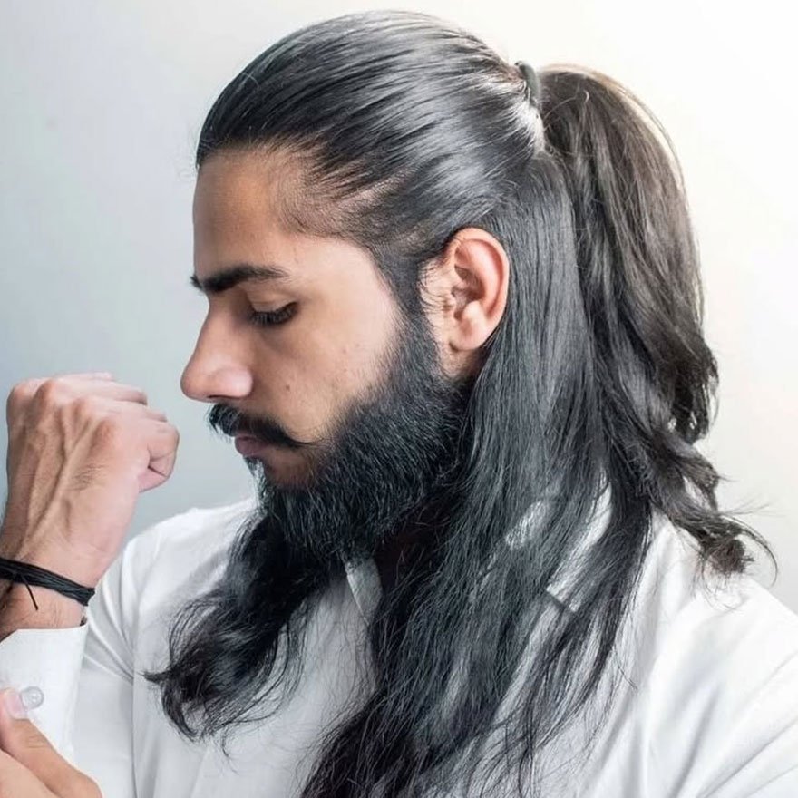 Man with long black hair styled in a sleek half ponytail, wearing a white shirt and showing a neatly groomed beard, looking down with a calm expression.