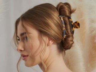 A woman with long, light brown hair styled in a chic claw clip updo, with loose face-framing strands and a tortoiseshell clip against a soft beige background.