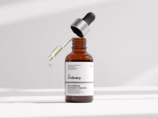 A realistic product photo of The Ordinary&rsquo;s Multi-Peptide Serum for Hair Density, featuring an amber bottle with a dropper releasing a droplet, placed on a clean white surface with soft light shadows.