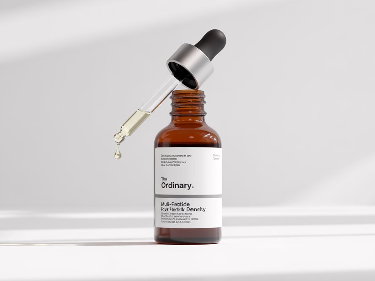 A realistic product photo of The Ordinary&rsquo;s Multi-Peptide Serum for Hair Density, featuring an amber bottle with a dropper releasing a droplet, placed on a clean white surface with soft light shadows.