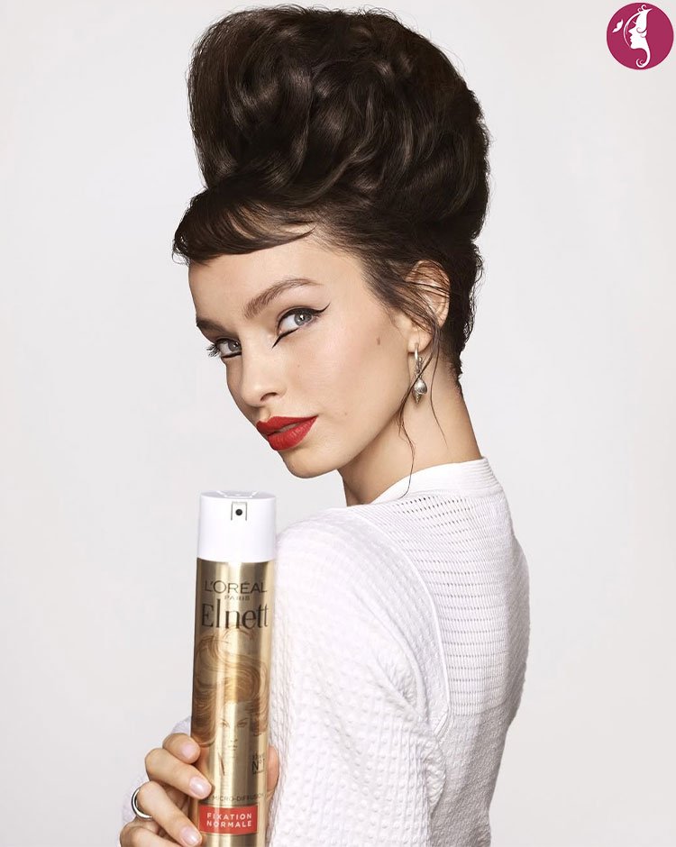 L'Or&eacute;al Paris Elnett Satin Hairspray held by a model with a voluminous hairstyle and bold makeup.