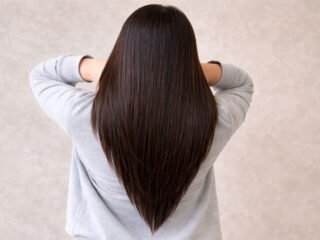 Back view of a woman with long, straight hair styled in a sharp V-cut, wearing a gray top against a neutral background.