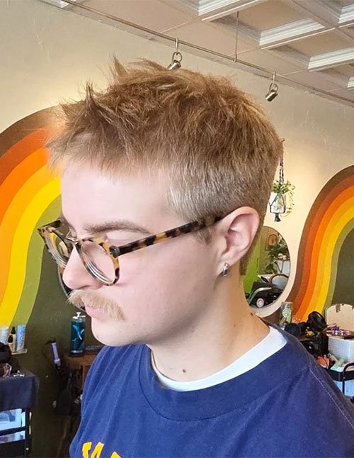 Side view of a person with a short 90s-style spiky gel haircut, featuring choppy textured spikes on top and closely trimmed sides, wearing glasses.