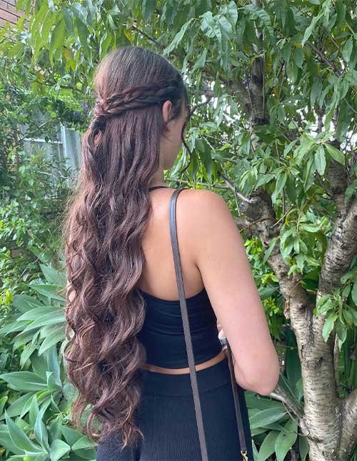 Back view of a woman wearing a braided formal half-up hairstyle with a delicate braid wrapped across the back and long loose waves.
