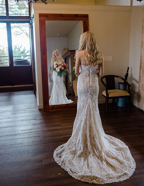Back view of a bride in a lace gown with medium hair worn down in loose, romantic boho waves, standing in front of a full-length mirror.