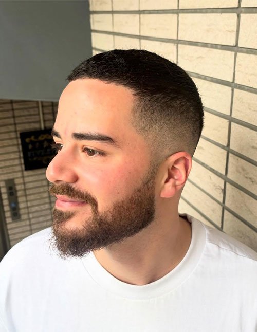 Angled side view of a man with a cropped buzz cut and a clean fade on the sides, paired with a full, neatly shaped beard.