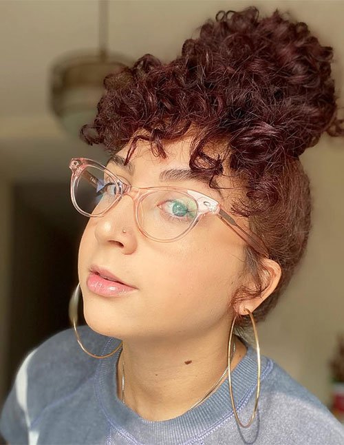 Close-up of a woman wearing glasses with a high curly bun and short curly bangs framing her forehead.