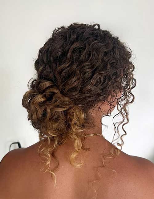 Side-back view of curly hair styled into a low curly chignon at the nape, with loose face-framing ringlets and caramel highlights.