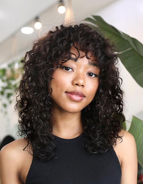 Woman with shoulder-length curly hair and soft curly bangs, photographed indoors with greenery in the background.