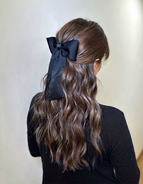 Back view of a girl with long, loose wavy hair styled half-up and secured with a large black bow.