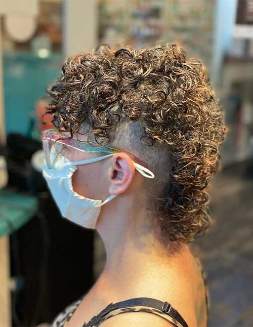 Side view of a curly mohawk hairstyle with shaved sides and back, featuring tight defined curls on top and down the center, with the person wearing a face mask.