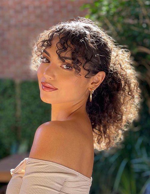Side-profile of a woman wearing a curly ponytail with loose face-framing curls and curly bangs.