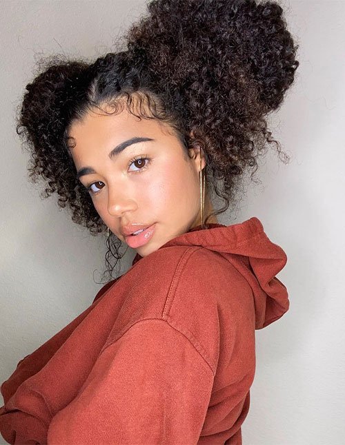 Woman posing in a hoodie with curly hair styled into two high double buns, with laid edges and soft curls framing the face.