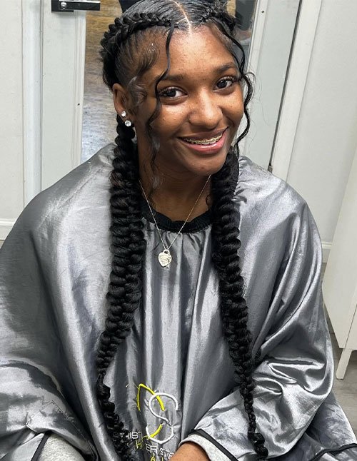 Smiling girl with a middle part and two long, thick raised braids, with curled tendrils framing her face.
