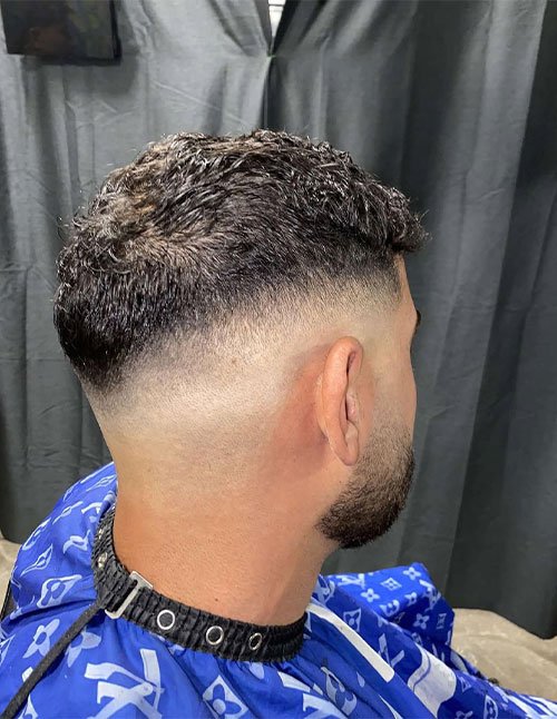 Back-side view of a man with a drop fade haircut that curves lower behind the ear, blending smoothly into short, textured curls on top.