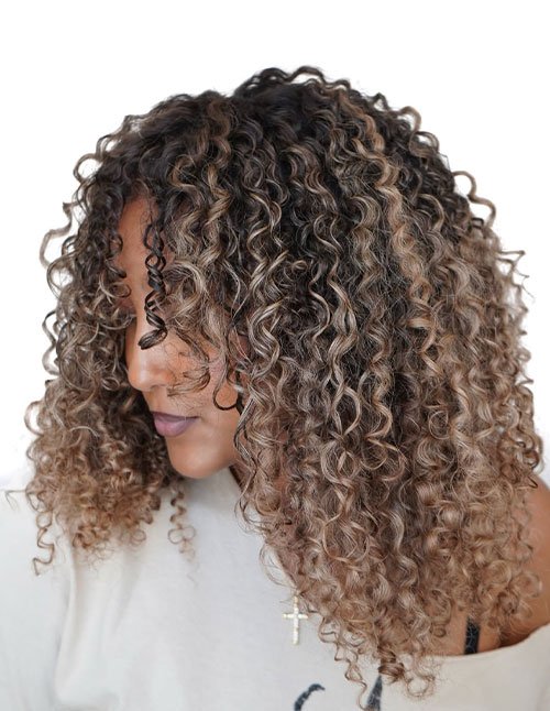 Side view of a girl with shoulder-length defined curls and balayage highlights, styled with a deep side part and face-framing ringlets.