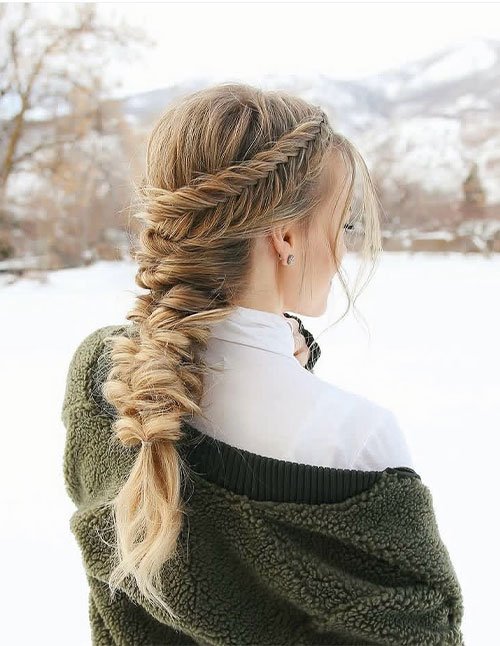 Back view of a woman wearing a loose fishtail braid with a side braid wrapped across the crown, standing in a snowy outdoor setting.