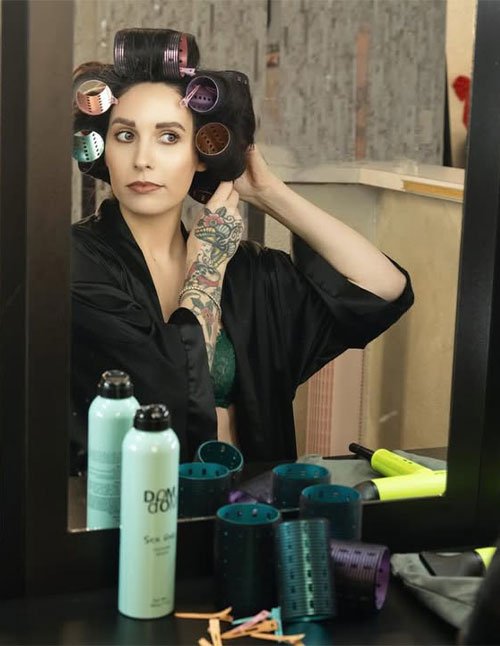 Woman looking in a mirror with several foam rollers set in her hair while getting ready at a vanity.