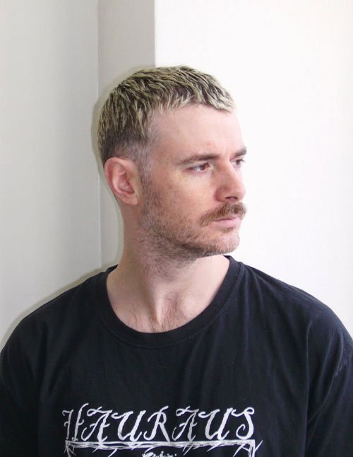 Man with a short cropped haircut featuring frosted tips, showing light blond highlights on the top while the sides remain darker and closely trimmed.