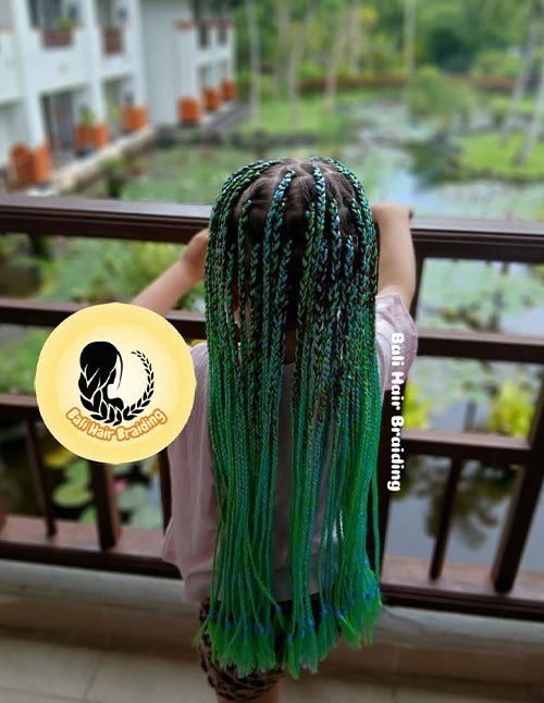 Child standing on a balcony with long box braids featuring green and teal color accents, styled neatly down the back.