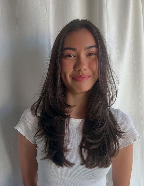 One woman with medium-to-long dark hair cut in soft layers, wearing a white top and smiling in front of light curtains.