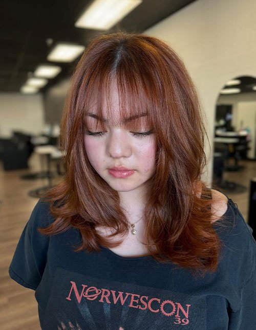 One woman with a medium-length layered haircut and soft curtain bangs, in a warm auburn tone, photographed indoors.