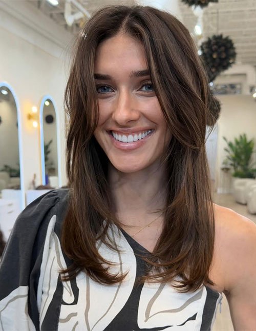 One woman with medium-length layered hair featuring soft, feathered ends and a center part, smiling indoors.