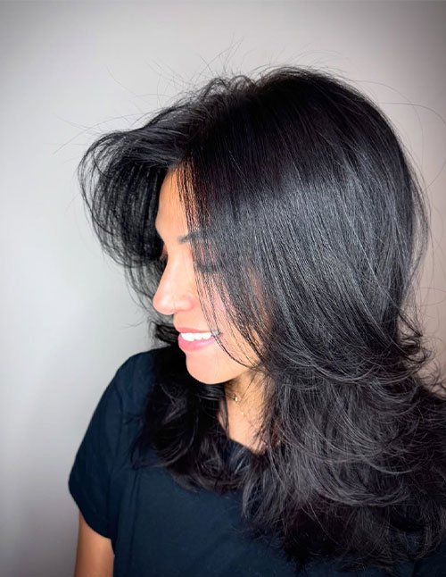Side view of one woman with medium-length black hair cut in layers, styled with a voluminous blowout and flipped ends.