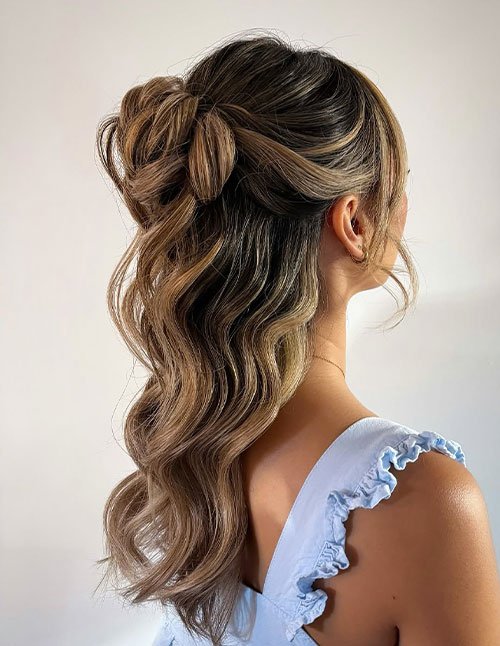 Side view of a woman with highlighted hair styled in a half-up bun, with the top section twisted into a loose bun and the rest of the hair left in soft, flowing waves.