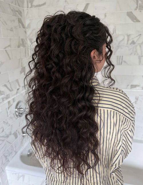 Back view of long dark curly hair styled half-up with a small bun on top and loose curls flowing down the back.