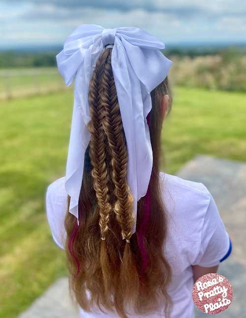 Back view of a girl with long hair styled in a half-up double fishtail braid, finished with a large white bow and loose hair flowing down.