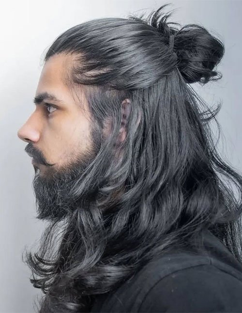 Side-profile of a man with long hair styled half-up into a small bun, with the rest left loose in soft waves.
