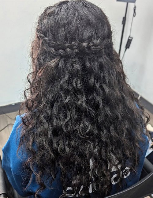 Back view of a woman with curly dark hair styled in a half-updo, featuring a braided band across the back with the rest of the curls left loose.