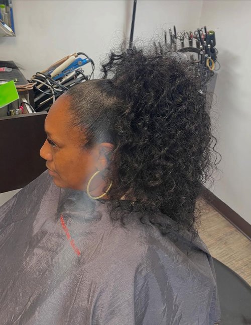 Side view of a woman with natural curly hair styled in a high ponytail, featuring sleek smoothed sides and full textured curls at the crown.