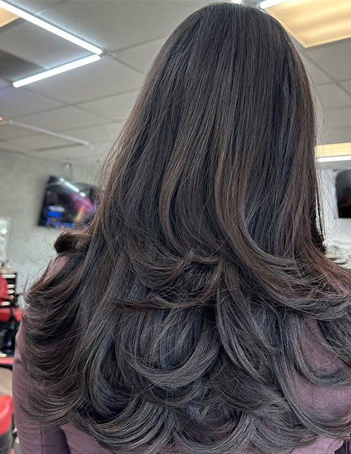 Back view of one woman with thick, medium-length dark hair cut in long layers, styled with a smooth blowout and curved ends.