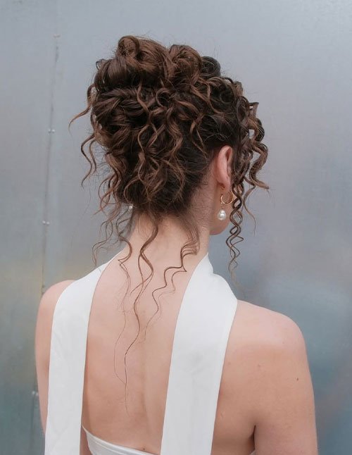 Back view of a woman with a loose curly updo, pinned curls for volume, and long curly tendrils left out, wearing a white dress and pearl drop earrings.