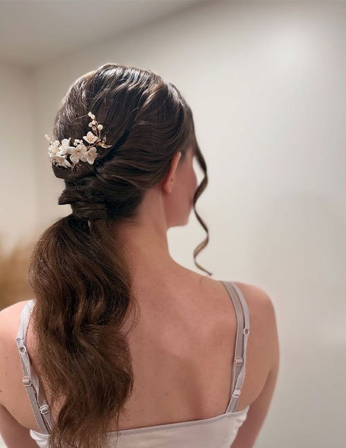 Back view of a woman with brown hair styled into a loose low ponytail, softly waved and secured with a floral hair accessory near the nape.