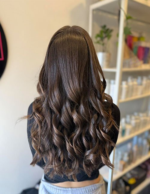 Back view of a girl with long brown hair styled in loose, natural-looking curls with soft shine.