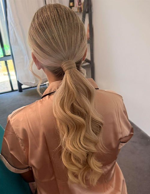 Back view of a low pony hairstyle with smooth blonde hair secured at the nape and finished with soft, loose waves.