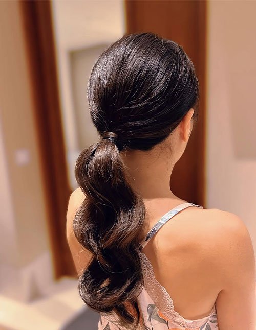 Woman wearing a low wrapped ponytail with smooth, sleek hair gathered at the nape and a section of hair wrapped around the base for a polished finish.