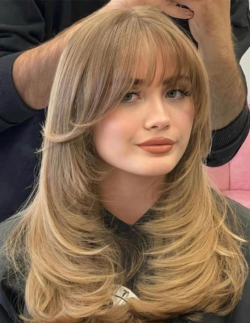 One woman with medium-length blonde hair styled in feathered layers and wispy bangs, photographed in a salon.