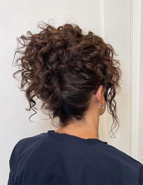 Back view of a woman with curly hair styled in a voluminous messy updo, featuring a textured curly bun with loose tendrils.
