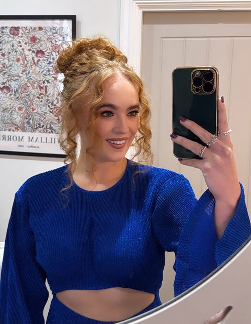 Woman taking a mirror selfie with a messy high bun and loose curled face-framing tendrils, wearing a royal blue long-sleeve crop top.