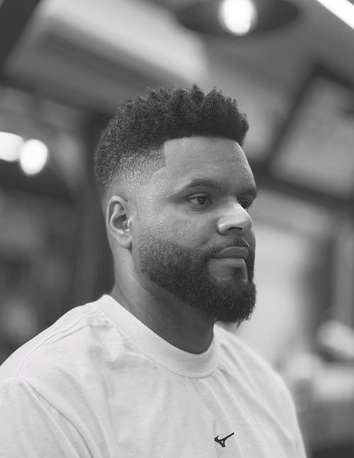 Side profile of a man with a mid skin fade freshcut, featuring a sharp line-up, clean bald fade around the sides, and a short textured curly top with a full beard.