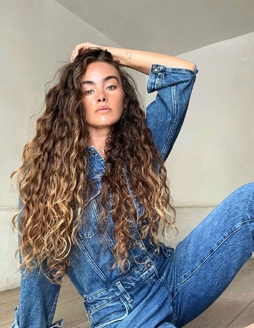 Woman with very long natural curls and subtle highlights, wearing a denim outfit.