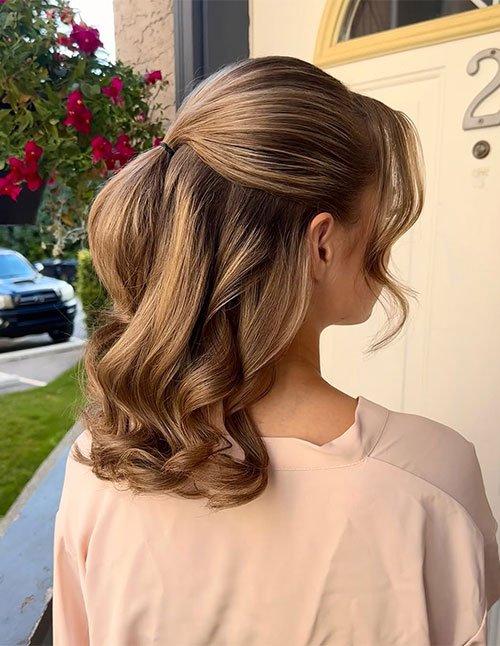 Back view of a half-up hairstyle with a smooth crown and loose, curled lengths, featuring warm blonde highlights and a polished, classic finish.