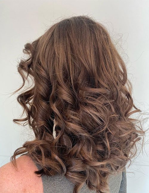 Back view of a girl with medium-long brown hair styled in voluminous, bouncy party curls.