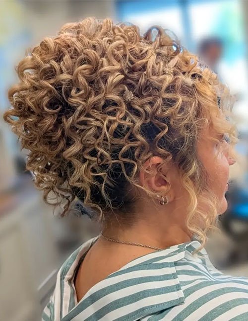 Side view of a woman with tight blonde curls styled into a pineapple updo, with loose face-framing ringlets.