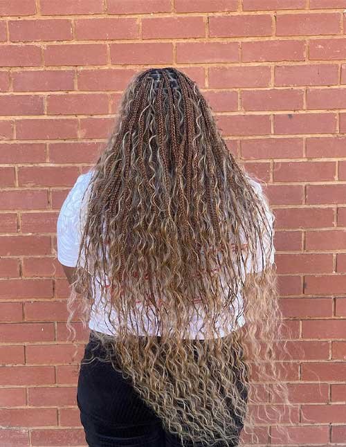 Long boho knotless braids with loose curls and a warm brown-to-honey ombré blend, shown from the back against a brick wall.