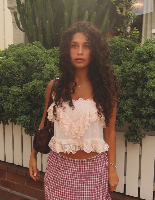 Woman with long, natural curly hair and a soft center part, standing outdoors in front of lush green plants.
